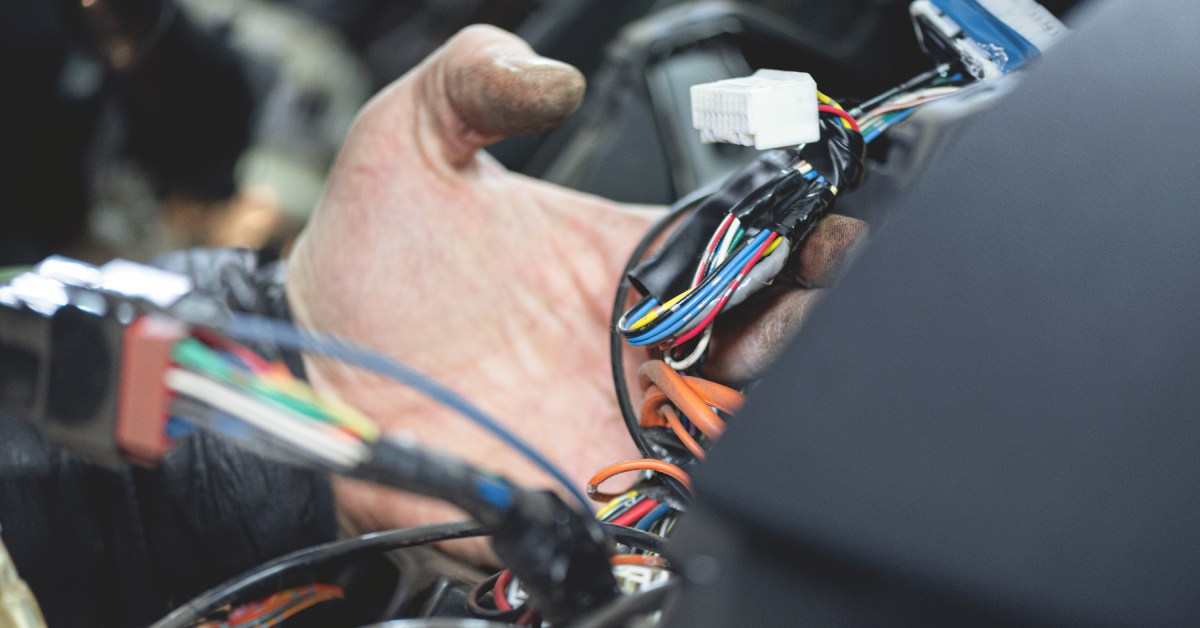 A hand is holding one of the wire systems in a vehicle. There are red, yellow, black, and blue wires tethered to connectors.