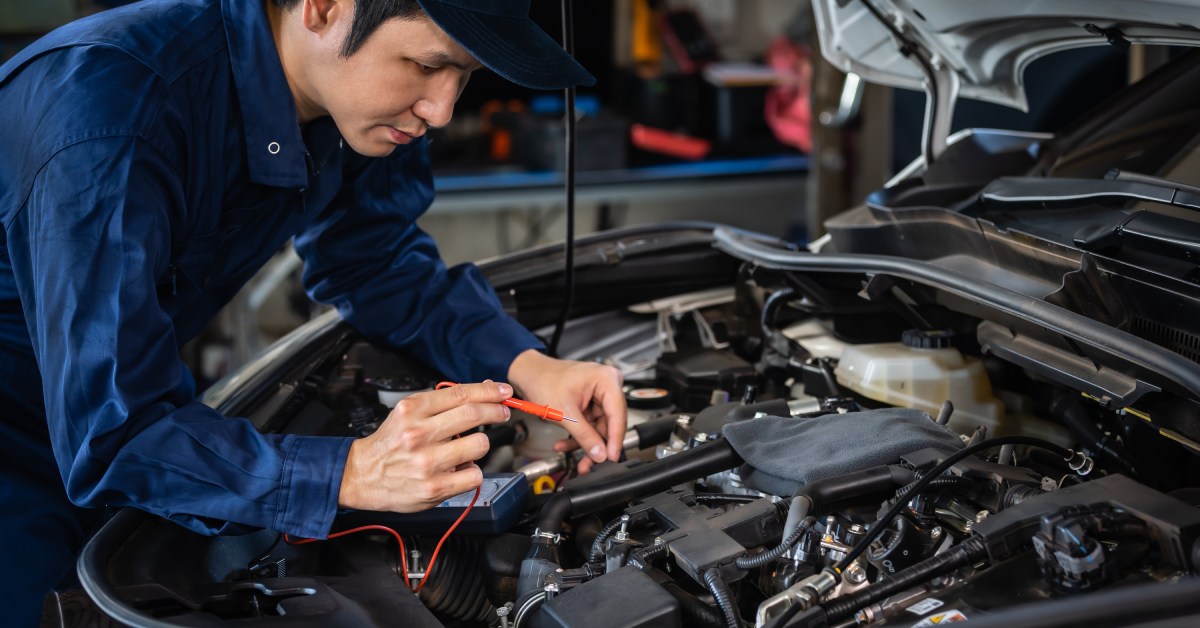 The hood of a car is open as a male mechanic works on the electrical systems inside of an automotive body shop.