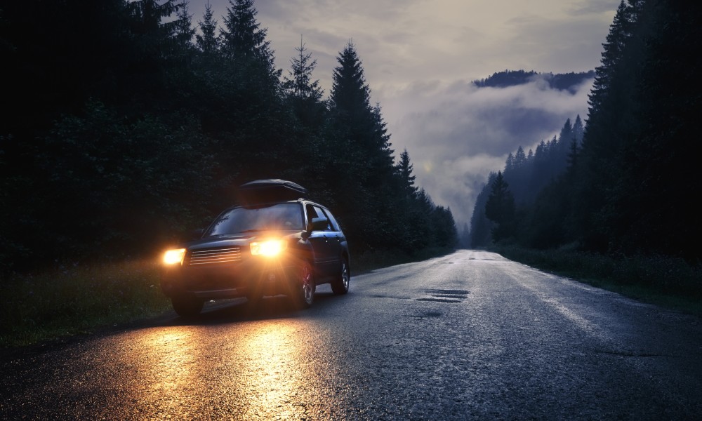 A mini-van driving with its headlights turned on as it travels down a dark road at dusk surrounded by pine trees.