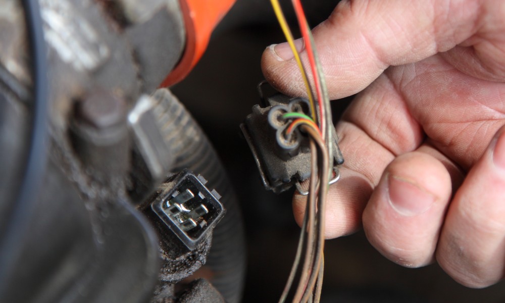 The hand of a mechanic holding a 4-pin automotive connector that’s connected to the colored wires of the vehicle.