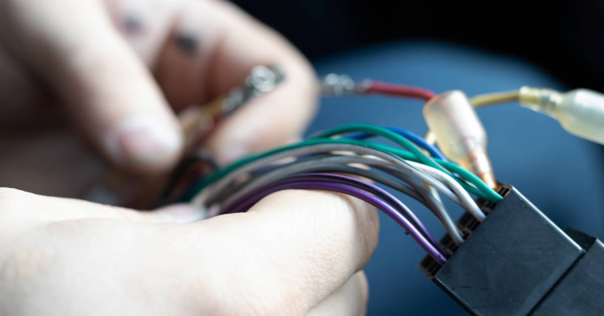 Two hands carefully hold color-coded wires that are connected to a black, rectangular electric connector.