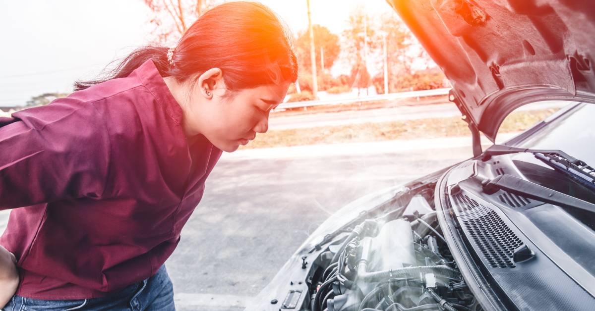 A woman in a red shirt and jeans stands outside with the hood of her car lifted as she inspects the smoking engine.