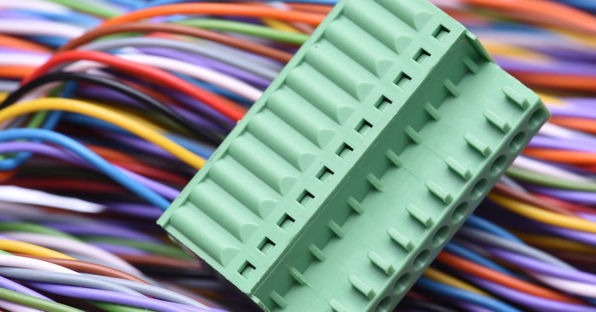 A rectangular, green connector with eleven pins connected to it lies on top of different colored electric wires.