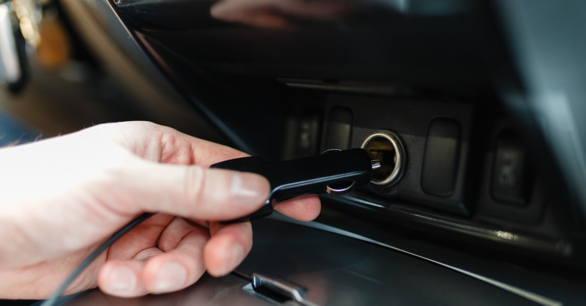 A hand holding an adaptor plug in front of the cigarette lighter to charge an electrical component inside the car.