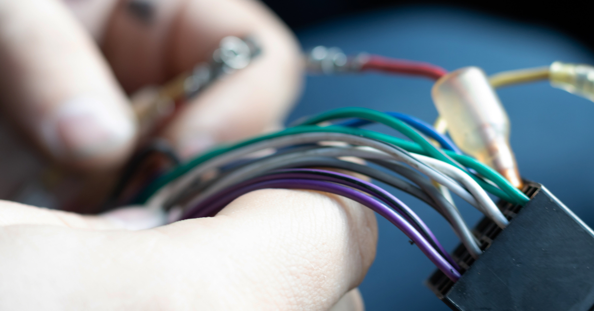 A pair of hands sorts and holds the connecting wires of a car's electrical system. There are purple, green, and white wires.