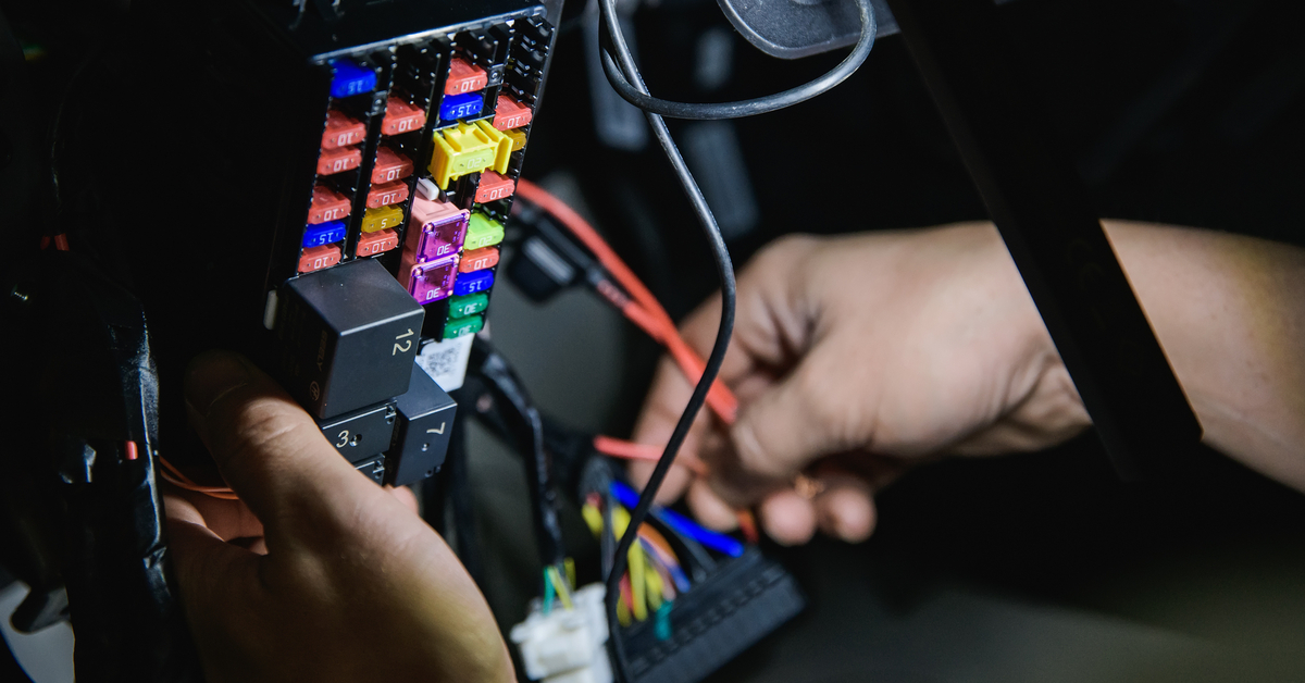 A pair of hands is running several colorful wires toward the fuses of a black automotive fuse box.