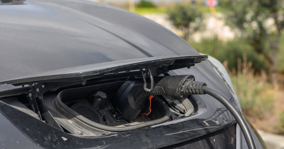 An electric charging cable is hooked up to the front engine of a small black sedan. Behind the car is a blurred visage of shrubbery.