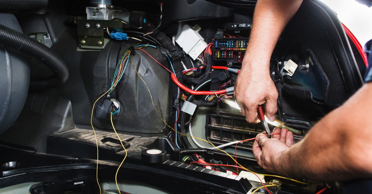 A pair of hands is working on a car's electrical system. Several colored wires are all leading to a connecting board.