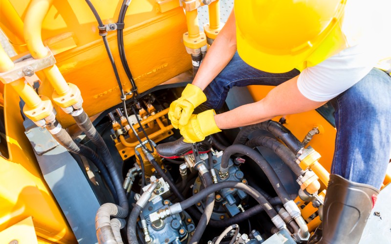  A construction worker performs maintenance on a machine's engine. Several wires and connectors are coming from the main block.