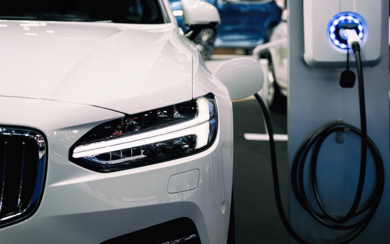 A white electric sedan hooked up to a charging station. The station has a faint blue circular light around the cable.