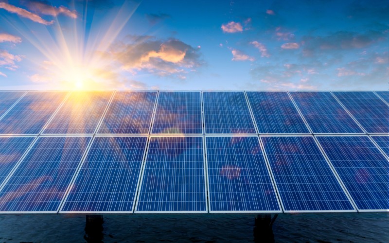 A wide range of solar panels aimed up toward the sky. The sun behind them creates a bright glare.