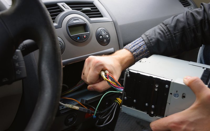 A pair of hands removing the infotainment system from a vehicle's dash. Several colorful wires lead into the empty hole.