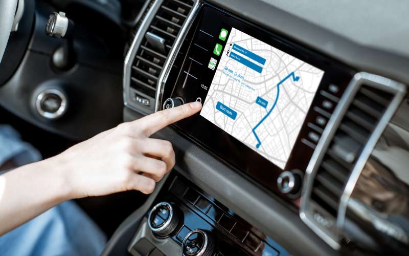 A hand is reaching out toward the touch screen in the center of a car. On the screen is a map revealing GPS instructions.