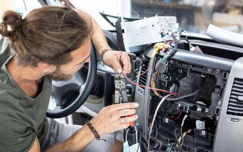 A man leaning into a car holds wires from the exposed open center console. He's using a pair of pliers on a red wire.