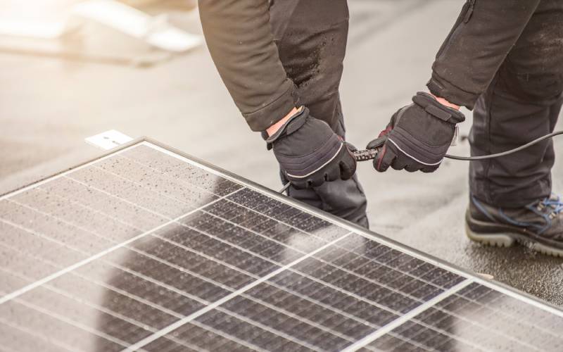 A person wearing black clothes and gloves connects a long black cable coming from a set of solar panels.