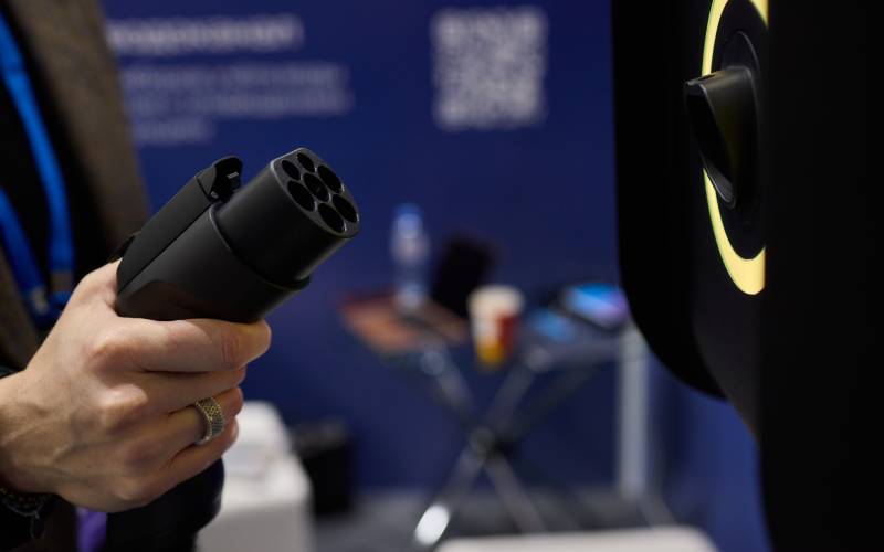 A hand holding a charging cable for an electric vehicle. The large black connector features seven round cavities.