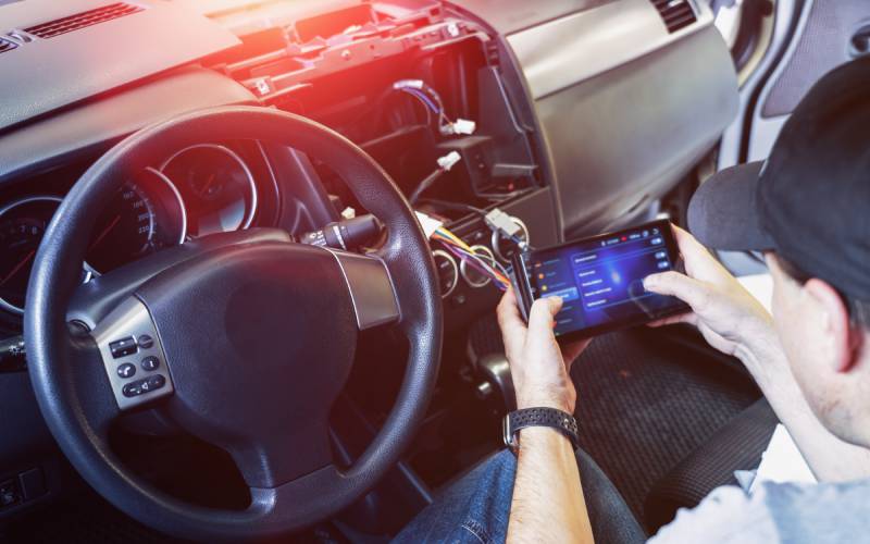 A man sitting in a car with an exposed center console. The wires and connectors run to a tablet with diagnostic information.