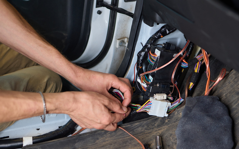 A close-up of a pair of hands working on a car’s electrical system. The car is white and the wires are multicolored.