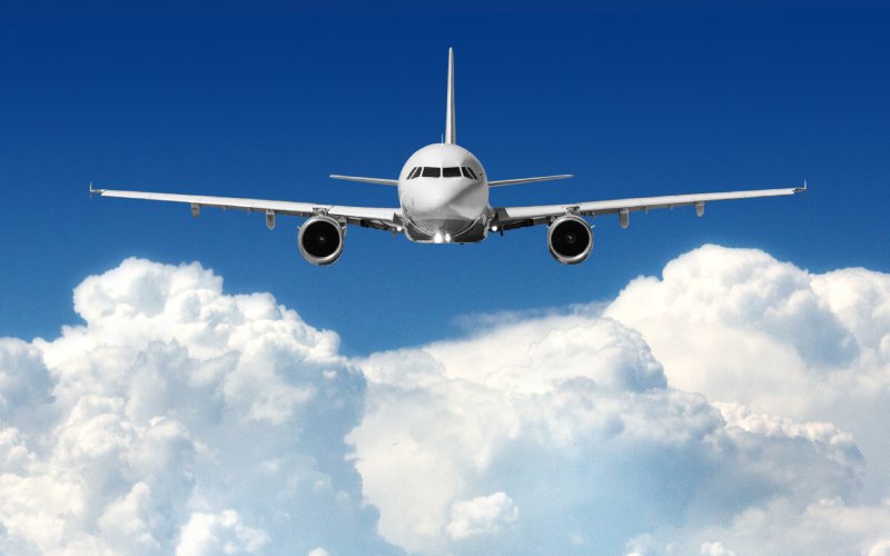 A white jet plane flies in the sky above fluffy, white clouds. The sky around it is a bright blue color.