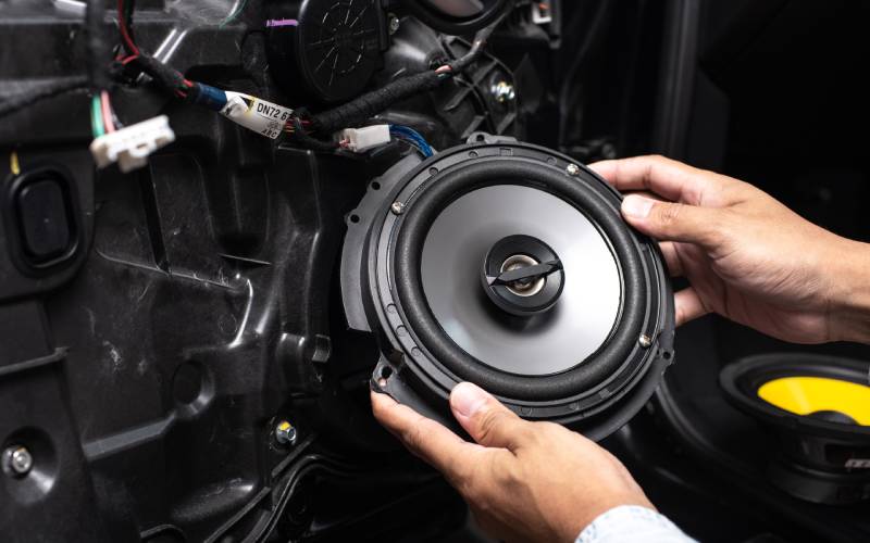 A pair of hands attaches a speaker to a car using multicolored wires connected with white connectors.
