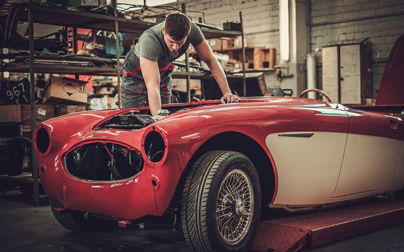 A man in a green t-shirt leaning over the open front of a red vintage car as he works on repairing it in an auto shop.