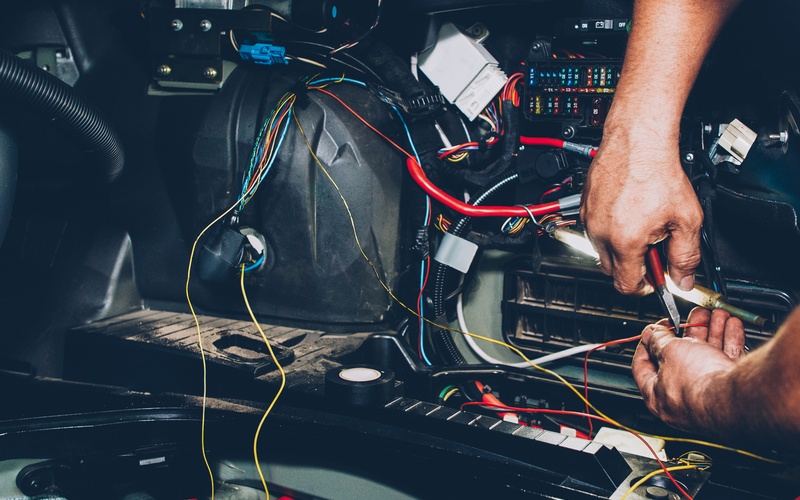 A pair of hands uses a tool on an orange wire that is part of a vehicle’s electrical system.