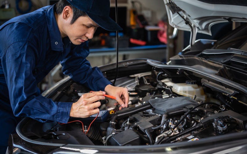  A mechanic wearing a blue jumpsuit and baseball cap checks the electrical system on a vehicle in a garage.