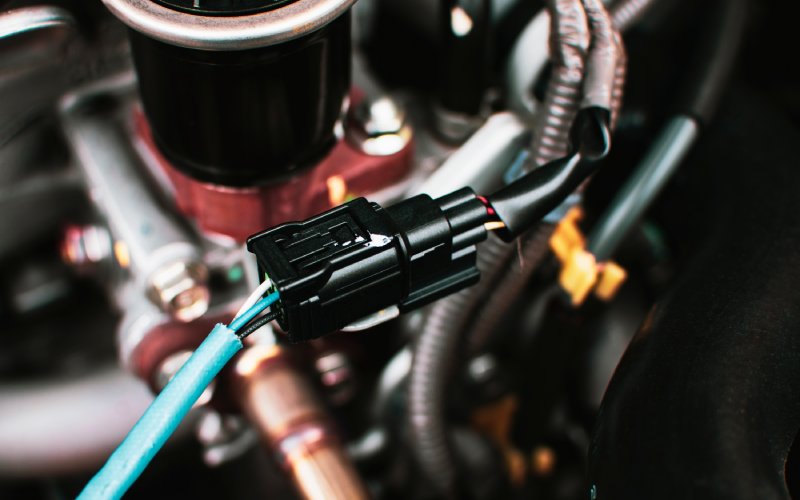 A light green wire plugged into a black automotive connector. A car engine is visible behind the connector and wire.