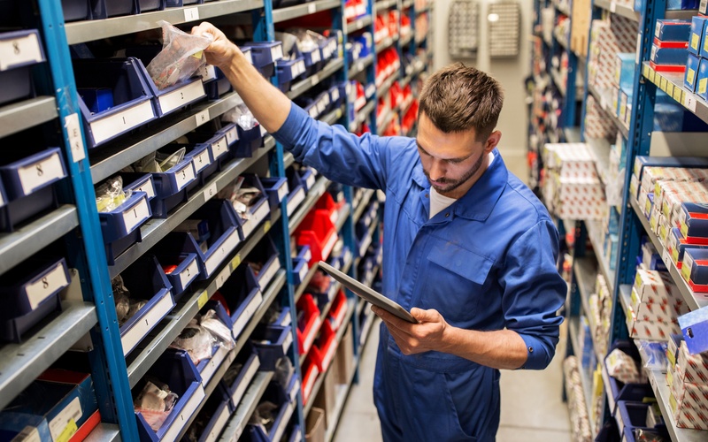 An employee wearing a blue shirt. The employee holds a tablet in one hand and pulls an item off a shelf with the other.