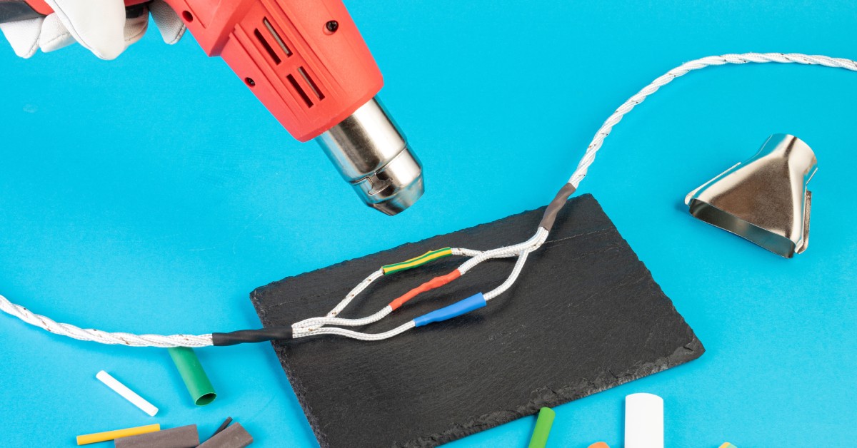 A hand wearing a white glove uses an orange heat gun on shrink wrap wire connectors on a blue surface.