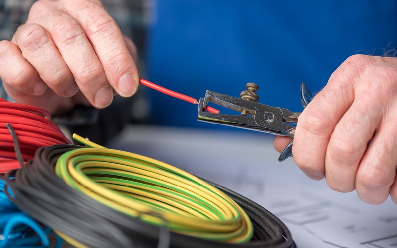 A close-up of an electrician wearing blue using a tool to strip a red wire near a pile of other coiled cables.