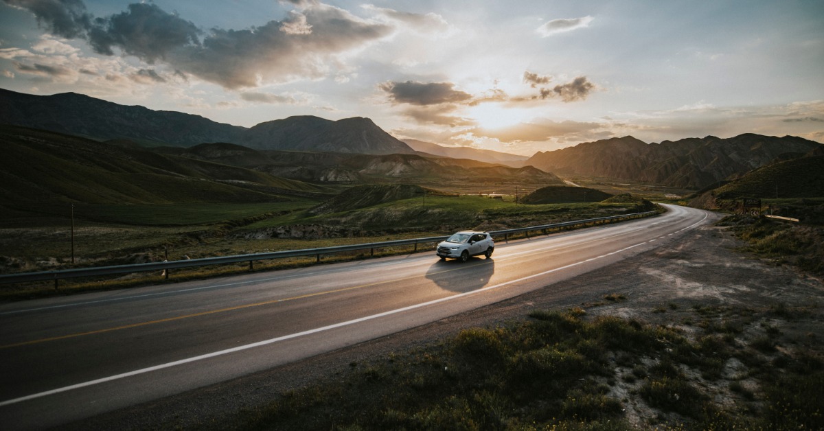A car driving on a long stretch of road. The sun is partially visible behind clouds and large mountains are nearby.