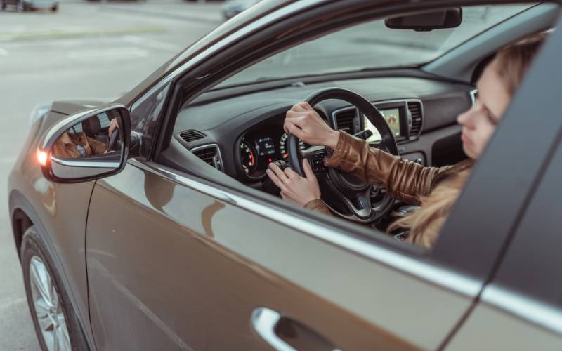 A person wearing a jacket and driving a brown car on the road. Both their hands are on the steering wheel.