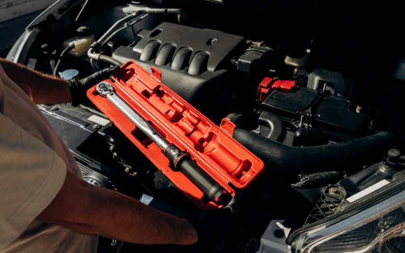 A mechanic wearing black gloves in front of a car while holding a red container with a torque wrench inside it.