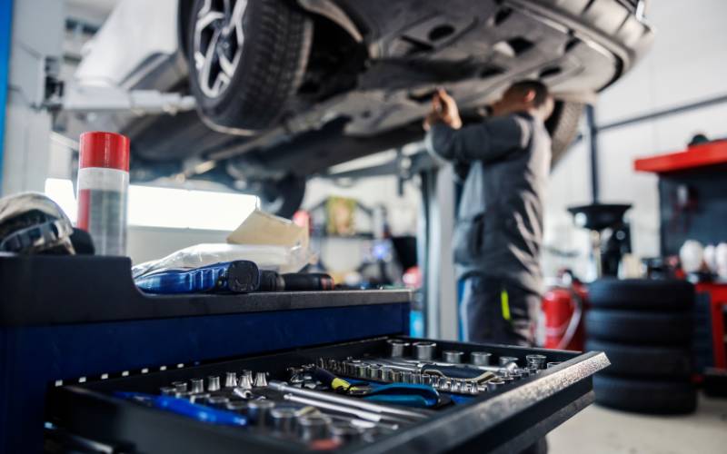 A mechanic working on the underside of a car in a repair shop. A toolbox full of tools is open nearby.