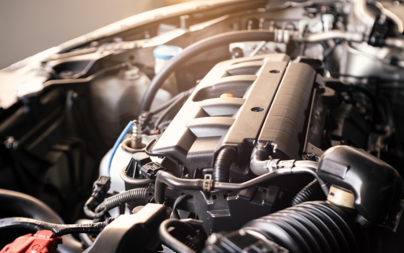 Sunlight shining on the open hood of a car. The open hood reveals a black engine with electrical wires and connectors.