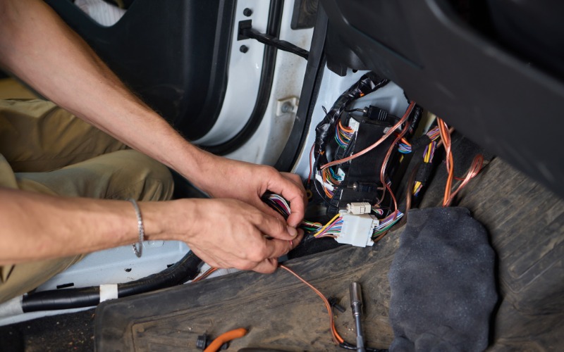 A mechanic repairing an automotive electrical system with white connectors. They work on the driver&rsquo;s side of the car.