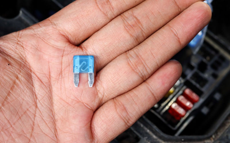 A person holding a small blue automotive fuse in front of a car. The fuse has two small metal connectors on it.