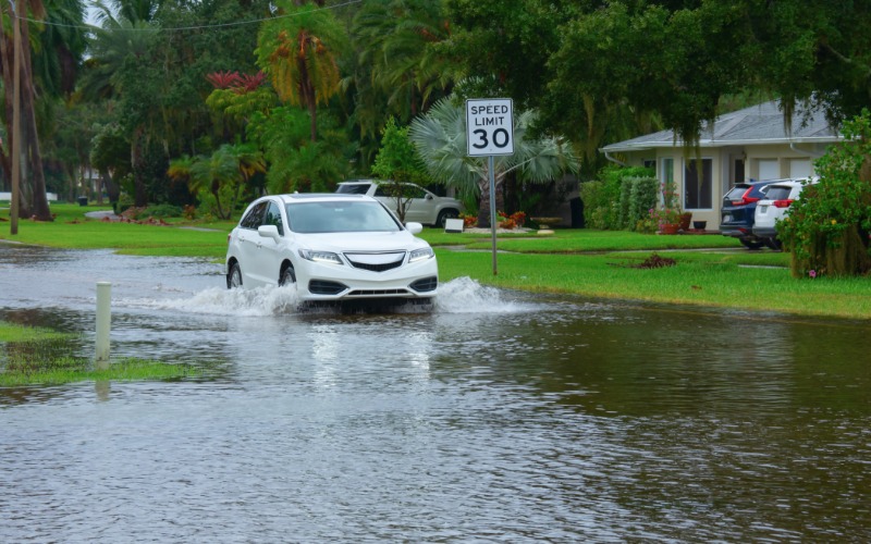 A small white car driving through a neighborhood street on a gray, gloomy day. The street is entirely flooded.