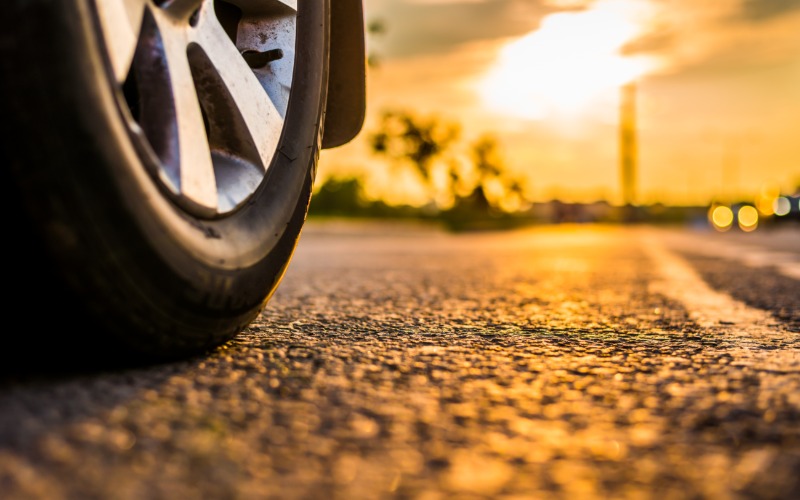 A car parked on the side of the road as other cars drive by it during the daytime. The sky is orange and sunny.