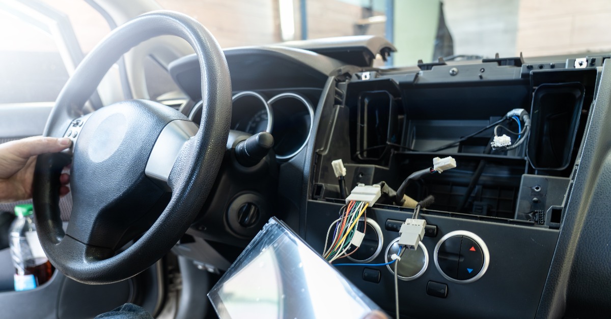 A person in a car with a new multi-media unit in their right hand. The dashboard is open to reveal wires and connectors.