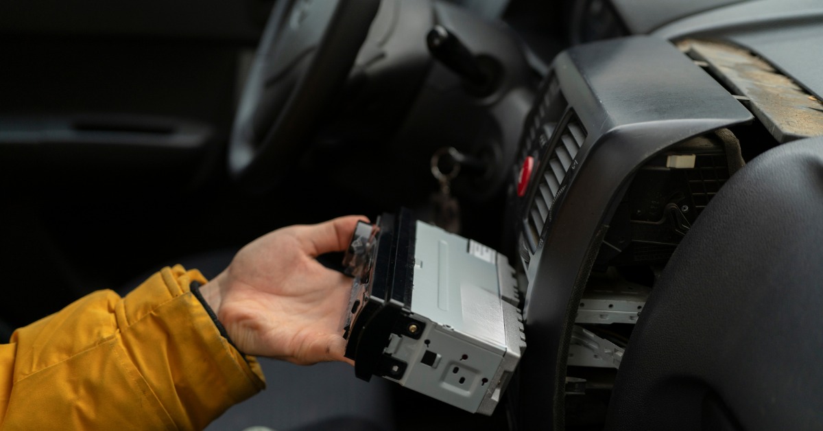 A person wearing a yellow jacket inserts a new radio into the dashboard inside a car with a black interior.