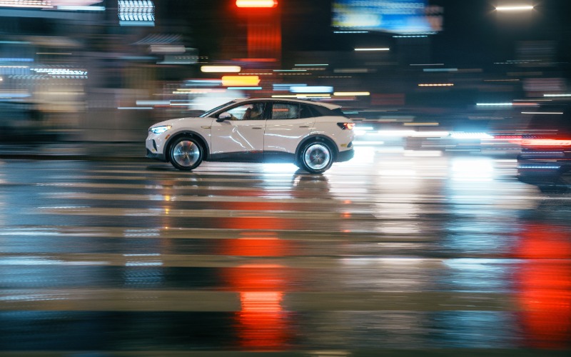 A sleek, white car driving past bright lights on a city street at night. The car drives over a crosswalk.