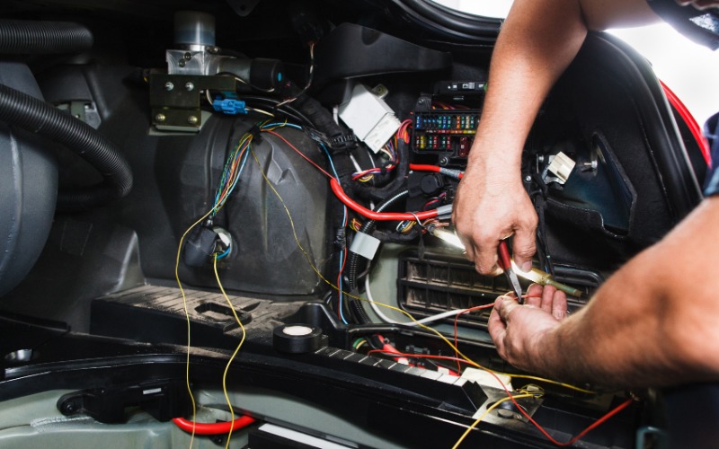 A mechanic using their bare hands to repair electrical connectors inside the side panel of a car trunk.