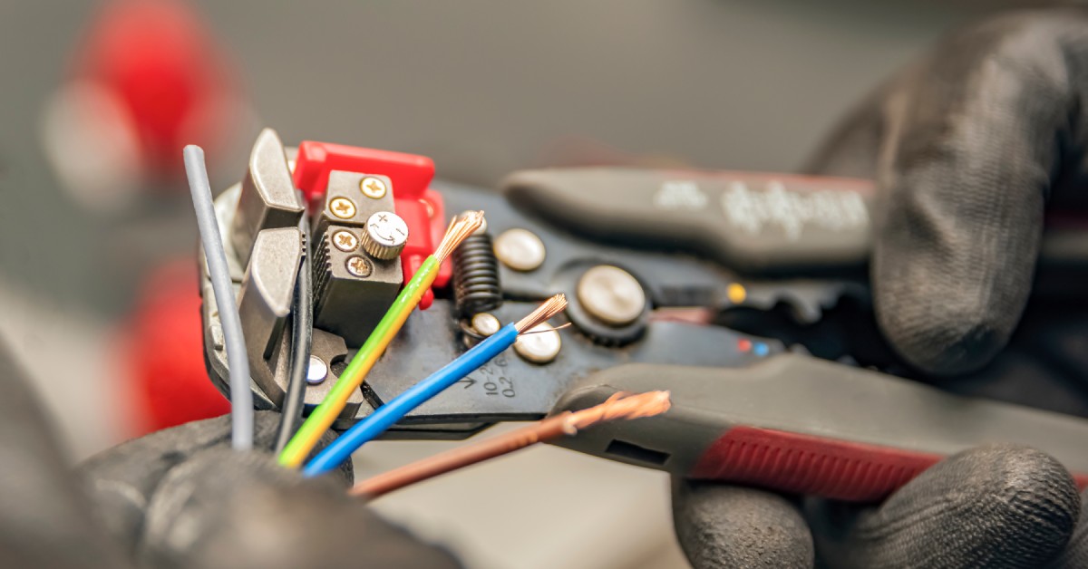 A person wearing thick work gloves and holding a wire stripper. The person uses the tool to work on a bundle of wires.