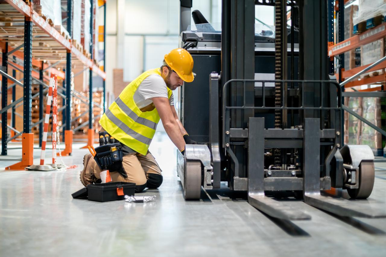 A mechanic wearing a hard hat and a toolbelt is leaning over a forklift in the middle of a warehouse to make repairs.