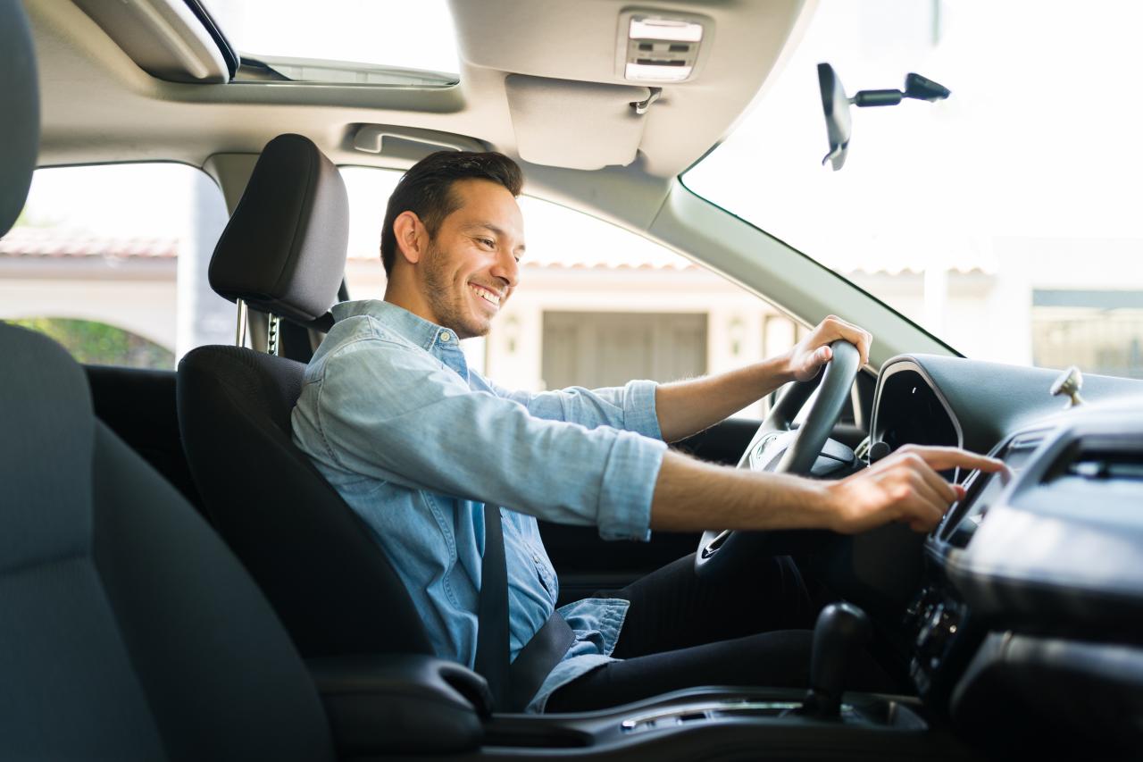 A driver smiling and holding their steering wheel with one hand and tapping a screen on their dashboard with the other.