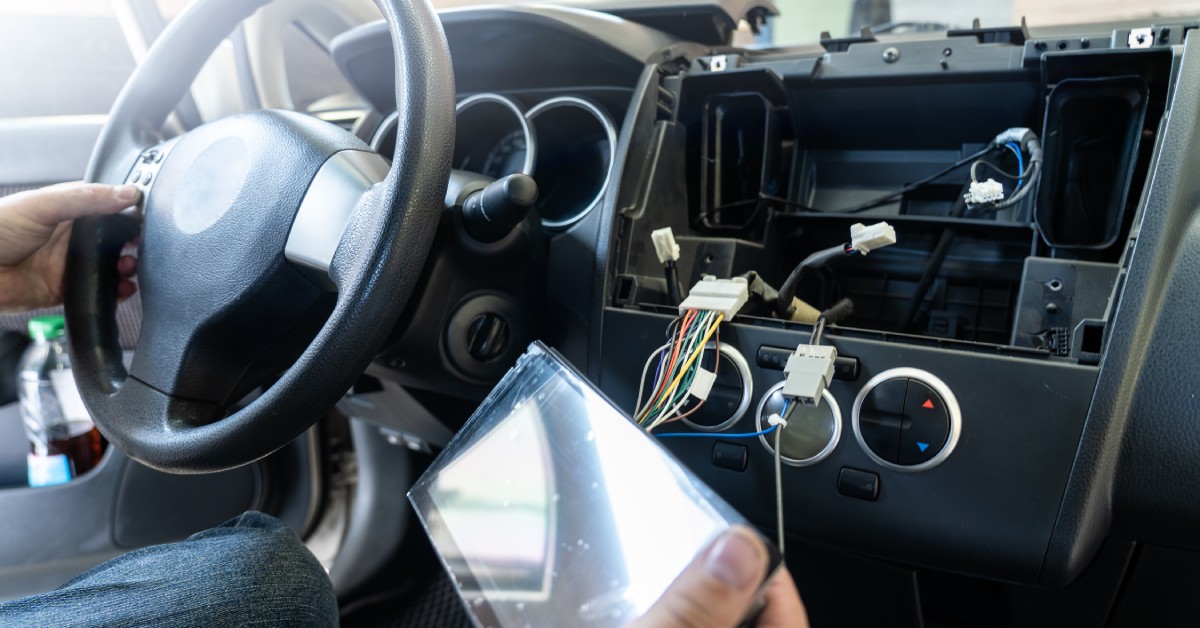 A person installing a monitor on the dashboard inside their car. Automotive connectors hang out the open dashboard.