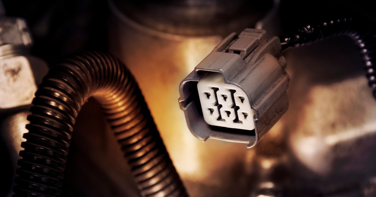 A six-pin automotive connector with a gray housing. The connector is placed next to a large black tube.
