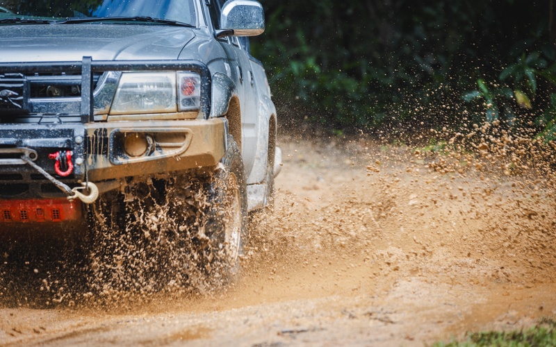 A large gray truck driving through a small puddle on an off-road trail. Mud splashes up from the puddle.